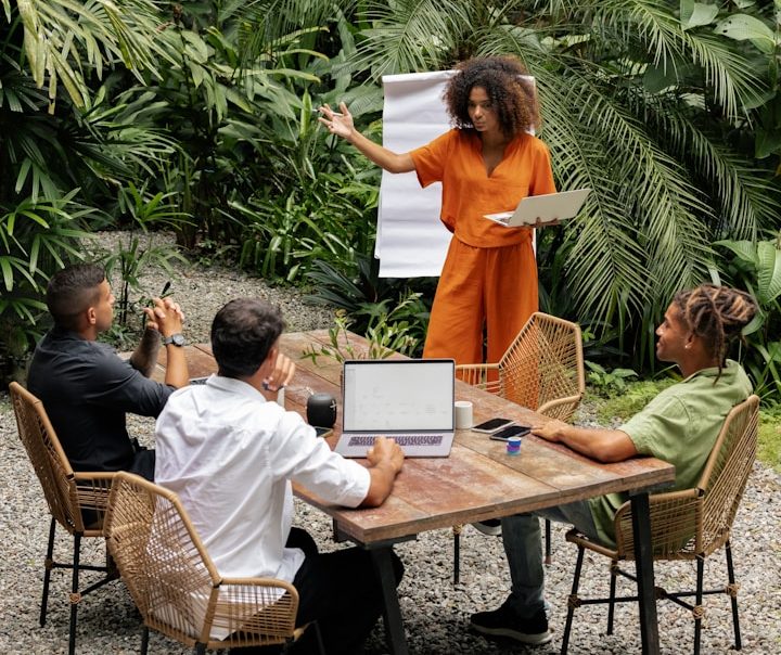 MasterClass & Formations en présentiel Une femme présente devant trois personnes lors d'une réunion en extérieur, entourée de verdure.